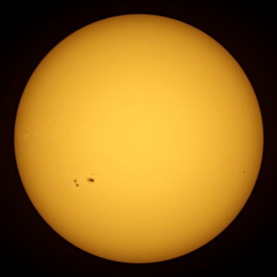 The Sun as a yellow-orange disc, darker towards the edge, with a group of dark sunspots lower left and a single small spot near the right edge